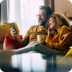A couple sit together at home on their couch, looking at a smartphone. 